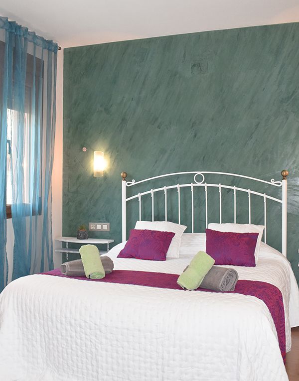 Habitación con cama de matrimonio. Tiene una de las paredes en tono verde y la colcha en blanco con cojines en púrpura. Cortinas en azul.